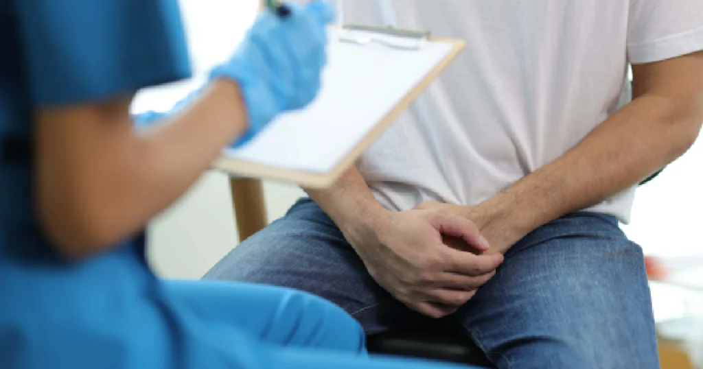 Erectile Dysfunction Treatment_ Man sitting with hands folded in lap during a medical consultation, as a healthcare professional in blue scrubs takes notes on a clipboard in Atlanta, GA.
